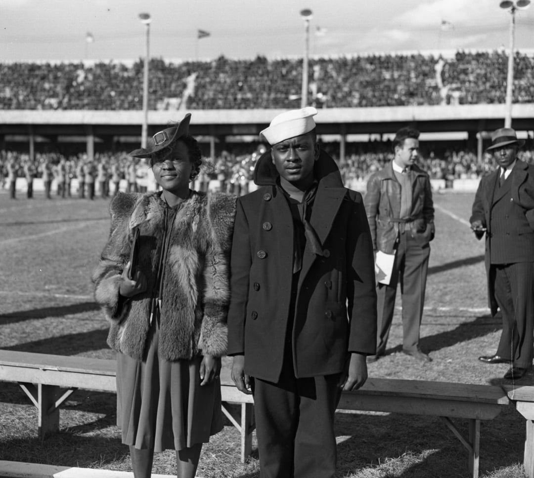 A woman in a fur coat and hat stands next to a man in a Navy uniform at a stadium event, with a large crowd and marching band visible in the background.