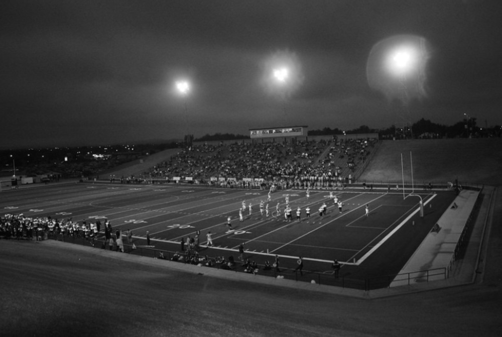 A black-and-white photo of a nighttime football game in a stadium, with players on the field, bright floodlights overhead, and spectators seated in the stands. The surrounding area is dark and the sky is cloudy.