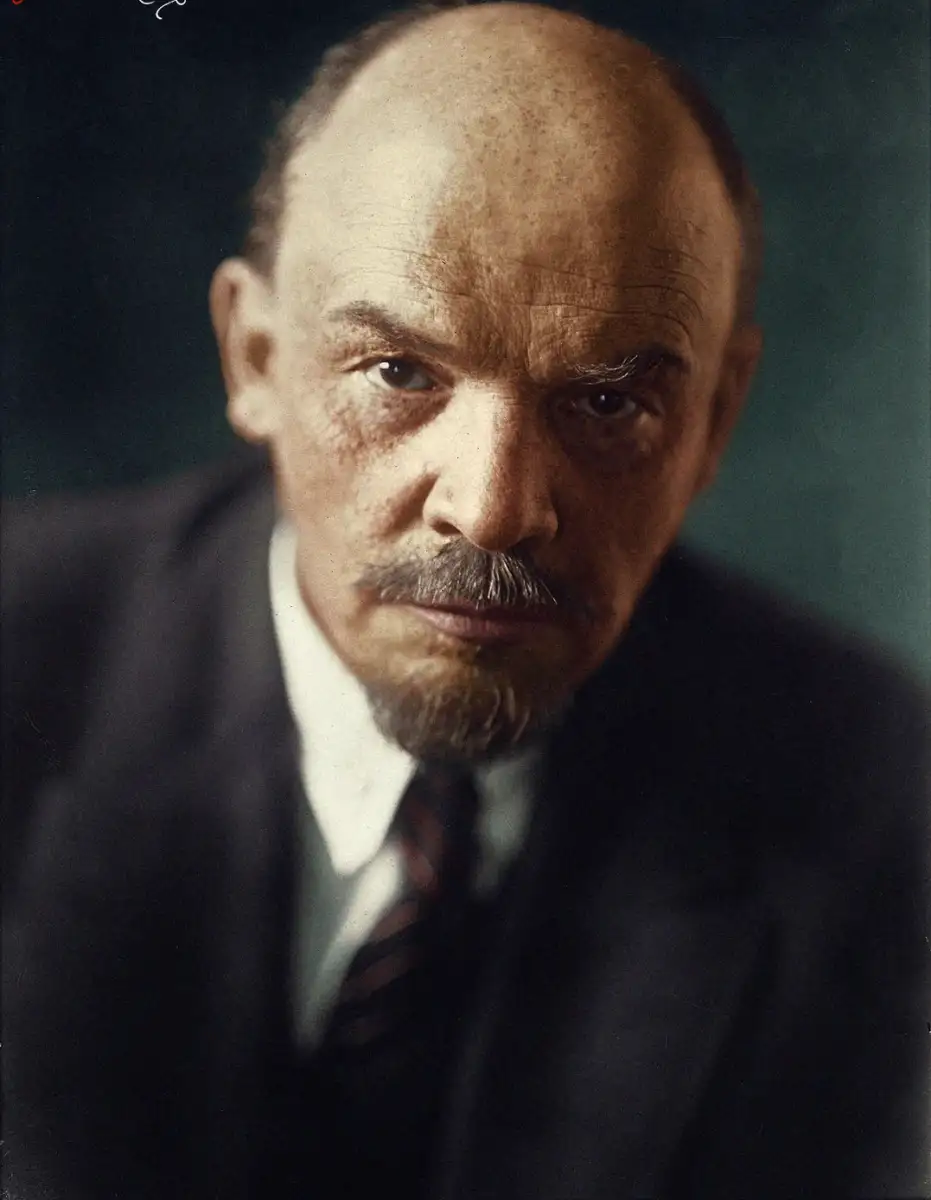 A bald man with a mustache and goatee, wearing a suit, white shirt, and striped tie, looks directly at the camera with a serious expression against a dark background.