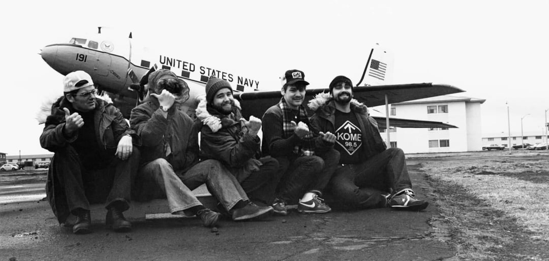 Five people in winter jackets sit on the ground smiling and giving thumbs up in front of a United States Navy airplane parked near a building. The photo is in black and white.