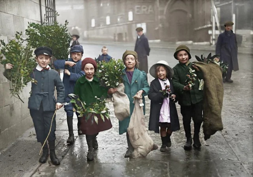 A group of smiling children in old-fashioned clothes walk down a city street on a rainy day, carrying greenery and sacks. Some wear hats and coats, and buildings are visible in the foggy background.