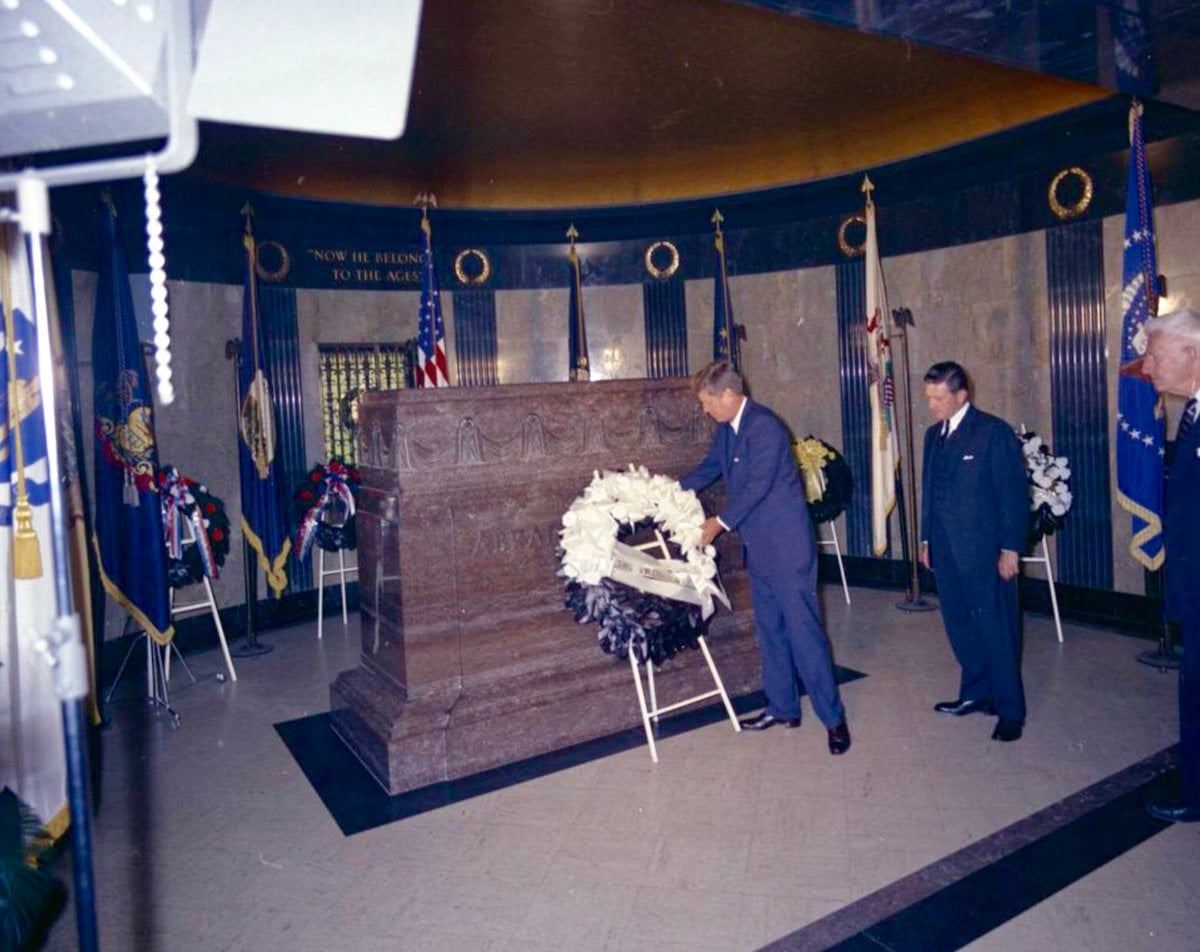 A man places a wreath in front of a large stone tomb inside a circular, dimly lit memorial room, with flags and other wreaths in the background and two men observing.