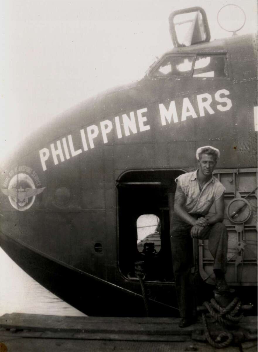 A man in a sleeveless shirt leans against the side hatch of a large aircraft named "PHILIPPINE MARS," which is docked near the water. The photo is black and white and appears vintage.
