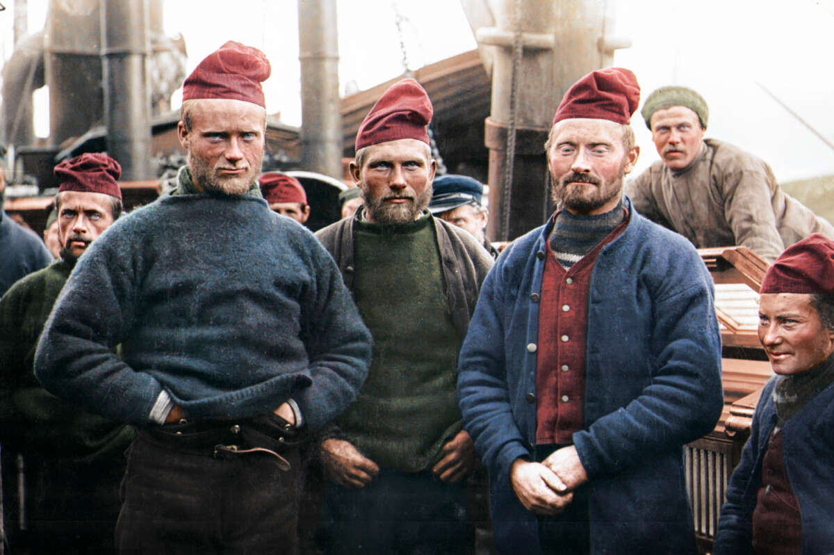 A group of men wearing red caps and heavy sweaters stand together on a ship’s deck, with others in similar attire in the background. The setting appears to be historic, possibly an early 20th-century expedition.