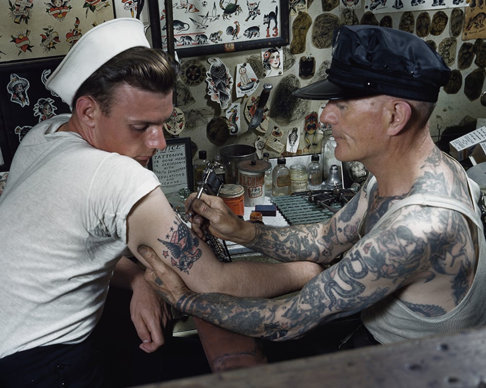 A tattoo artist with tattoos on his arms inks a design onto the upper arm of a man in a sailor uniform. The room has tattoo designs on the walls and various tattoo equipment on the counter.
