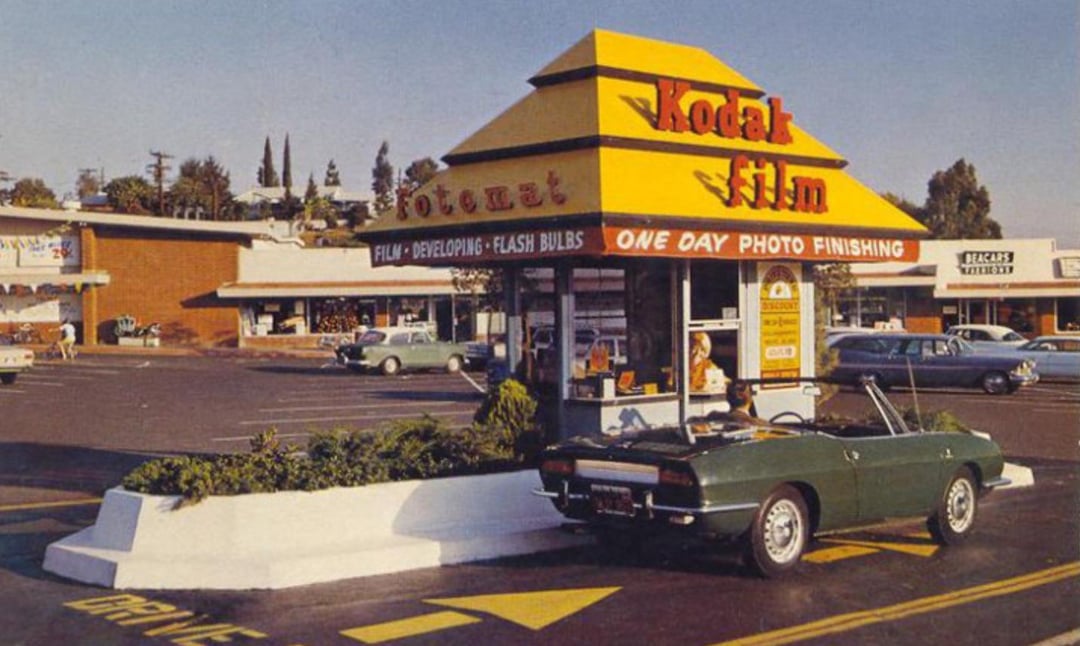 A vintage Kodak photo kiosk stands in a parking lot with a yellow roof and red signage. A green convertible car is parked beside it. Shops and parked cars are visible in the background under a clear sky.