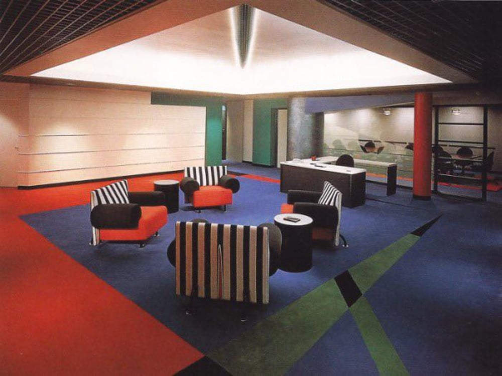 A modern office lobby with colorful, geometric carpet, striped and solid-colored armchairs, small round tables, and a large desk; glass wall reveals a conference room in the background.