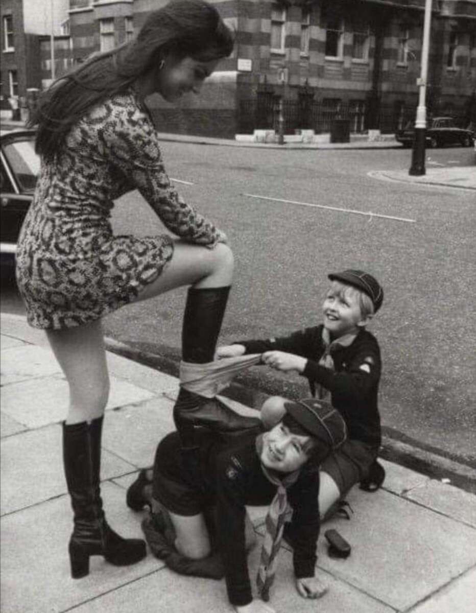 A woman in a patterned dress and knee-high boots stands on a sidewalk with her foot on a kneeling boy. Another boy, both dressed in scout uniforms, wraps a scarf around her leg, all three smiling and appearing playful.