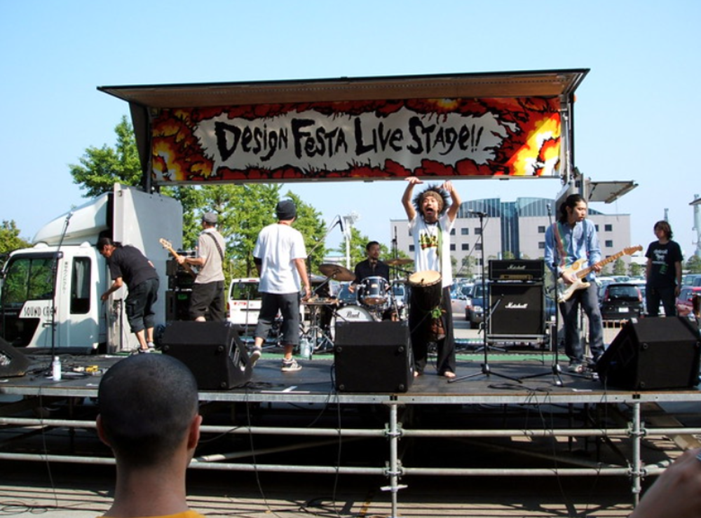 A band performs on an outdoor stage labeled "Design Festa Live Stage." Several musicians play instruments while one person sings energetically. Audience members watch from the foreground under a clear sky.
