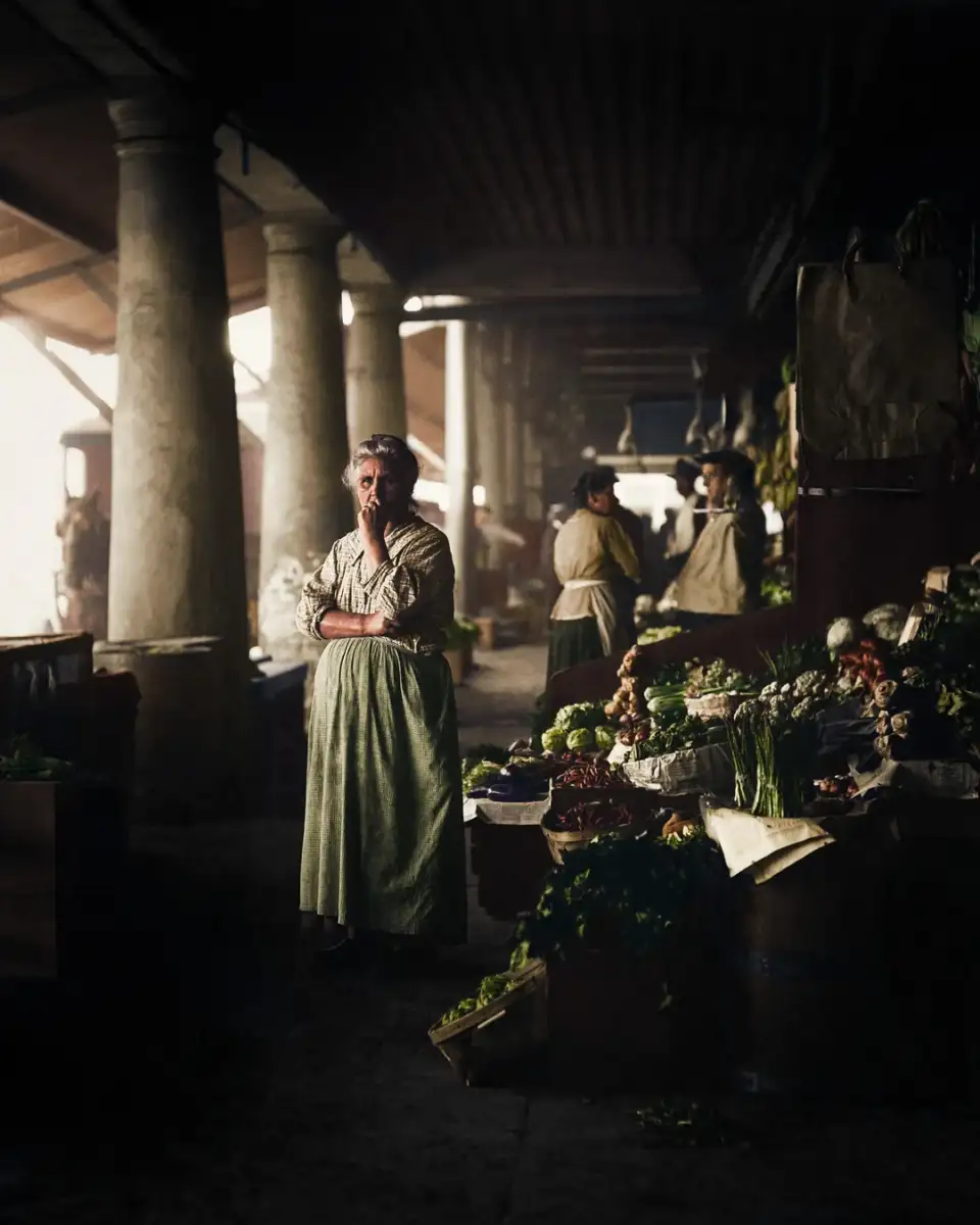 A woman in an apron stands with arms crossed beside a vegetable stall in a dimly lit, old market with high columns; other women work at stalls in the background.