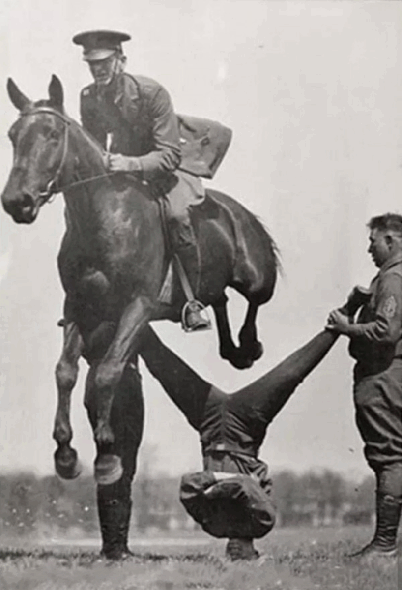 A soldier on horseback jumps over another soldier who is doing a handstand, while a third soldier holds the handstand performer's legs. The scene appears to be part of a military training or demonstration.
