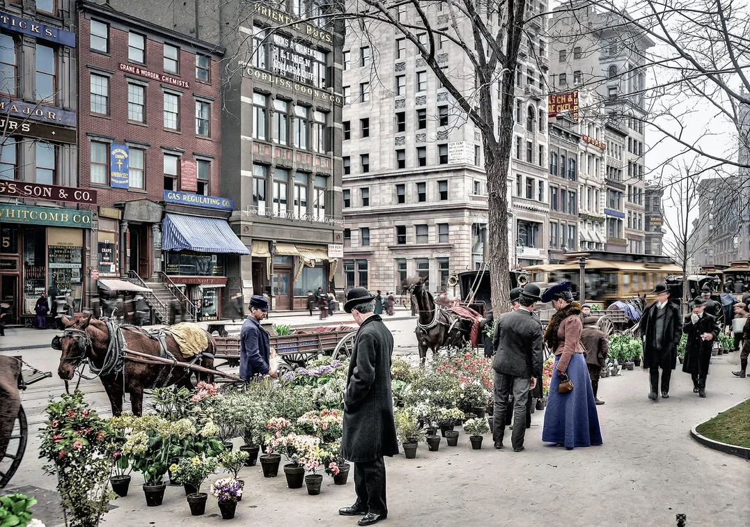 People in early 20th-century attire shop for potted plants and flowers at an outdoor market on a city sidewalk, with horse-drawn carriages and tall buildings in the background.