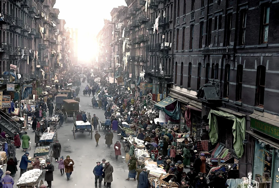 A busy street market scene in a historic city, crowded with people, street vendors, carts, and stalls. Tall buildings line both sides of the street, and the sun shines brightly in the distance.