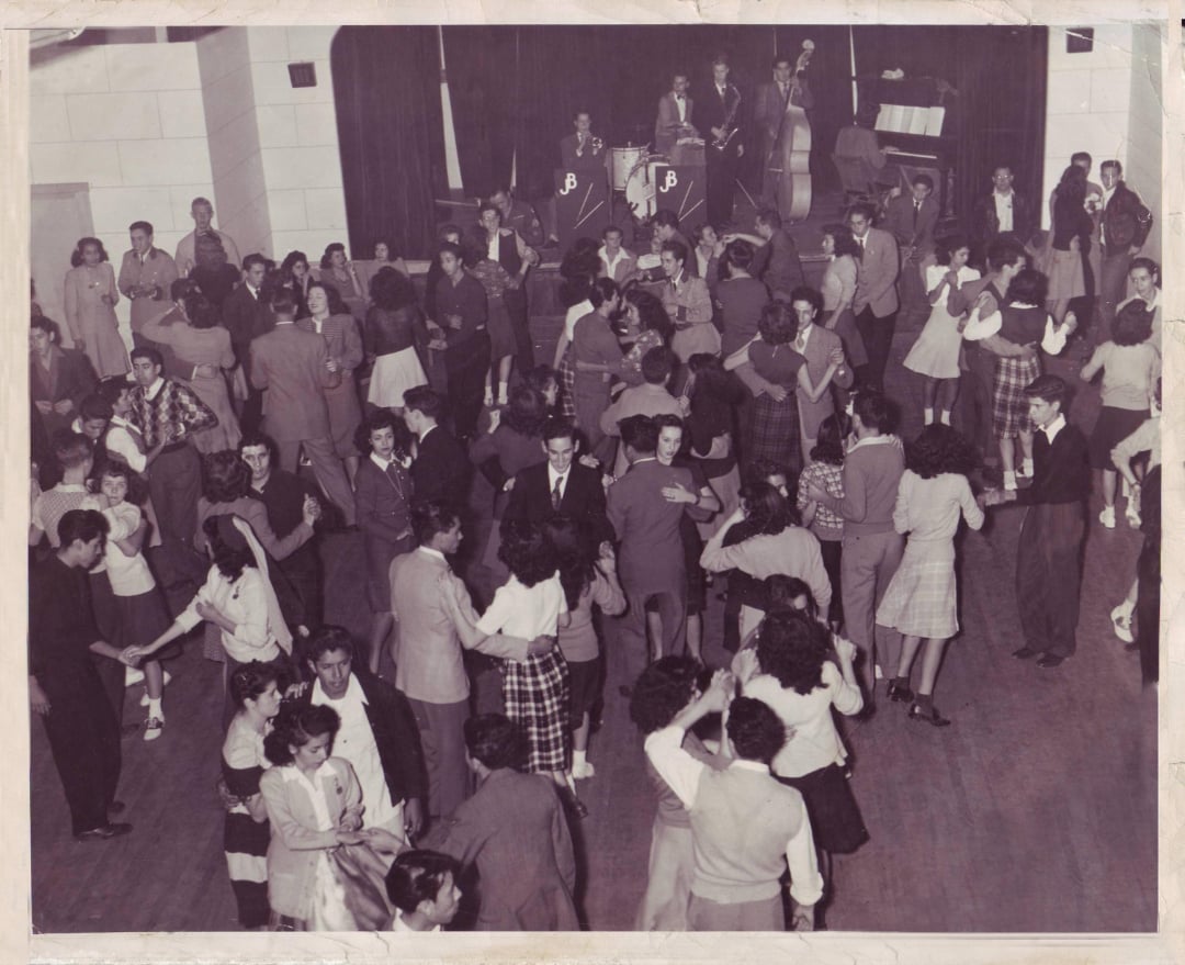 A large group of people dance in pairs in a hall with a wooden floor, while a live band plays onstage in the background. The scene appears to be from a mid-20th century dance event or social gathering.