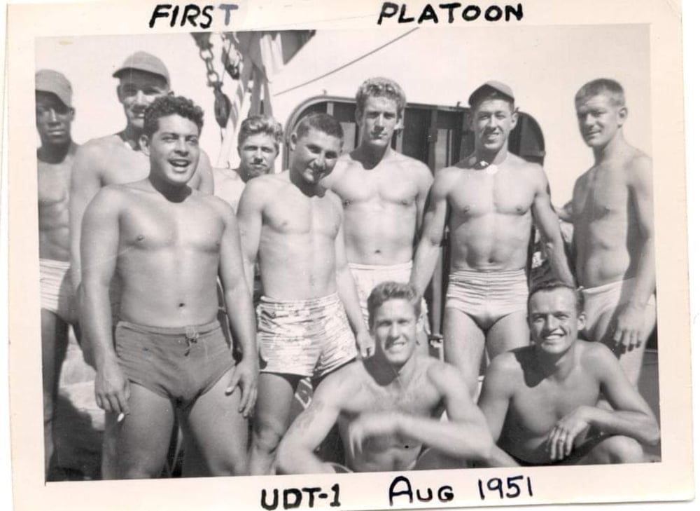A black-and-white photo of nine men in swim trunks posing outdoors, smiling. "FIRST PLATOON UDT-1 Aug 1951" is handwritten on the photo. A building is visible in the background.