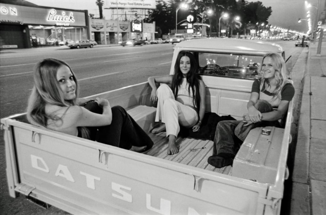 Three young women sit and smile in the back of a Datsun pickup truck parked on a city street at night, with bright lights, cars, and storefronts visible in the background.