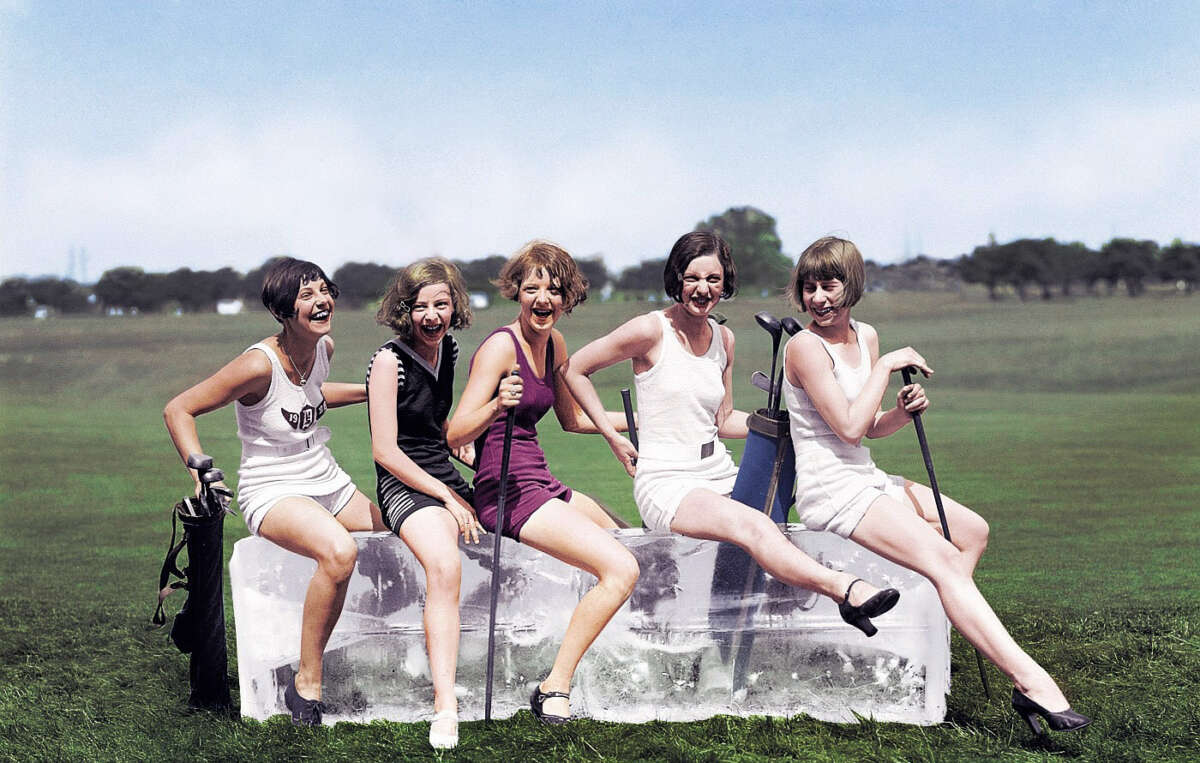 Five women in 1920s-style swimwear sit and laugh on large blocks of ice outdoors, each holding a golf club, with a grassy field and trees in the background.