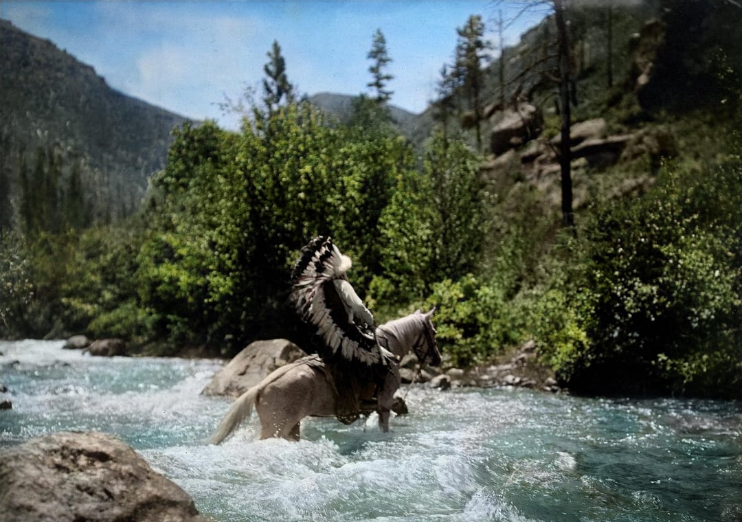 A person wearing a feathered headdress rides a white horse through a flowing river, surrounded by lush green trees and rocky hills under a blue sky.