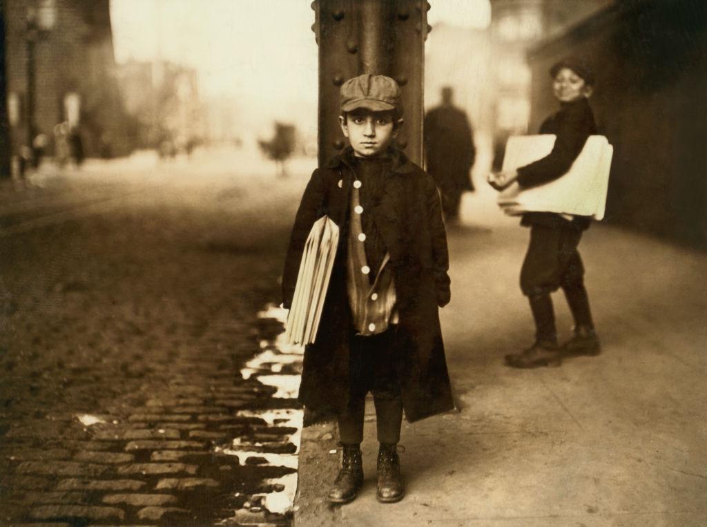 A young newsboy stands on a cobblestone street holding newspapers, wearing a cap and coat. Another boy with newspapers stands in the background. The scene is in sepia tones and appears to be from the early 1900s.