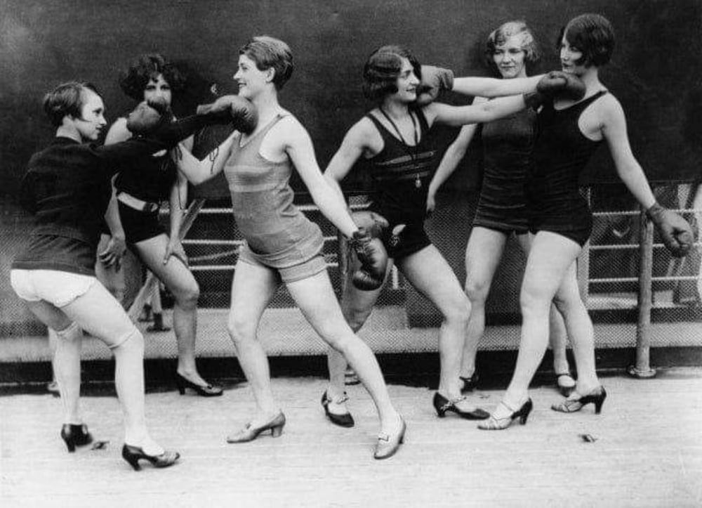 Six women wearing swimsuits, boxing gloves, and heeled shoes playfully spar with each other in a vintage black-and-white photo, smiling and posing on a wooden floor with a rope barrier behind them.