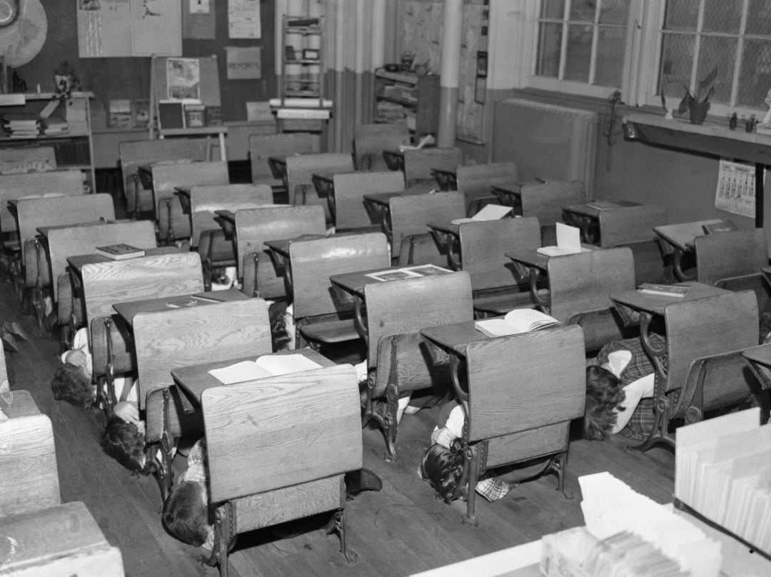 Black-and-white photo of children hiding under their wooden school desks during a drill in a classroom, with books left open on the desks and classroom materials on the walls.