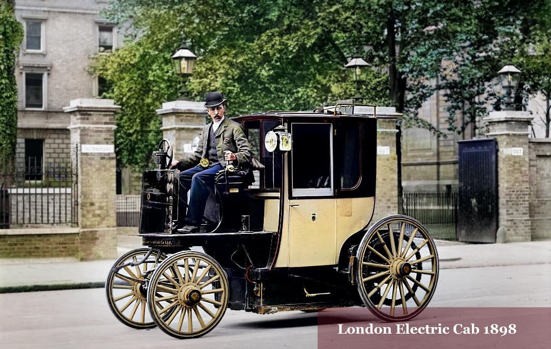 A man in vintage attire drives a historic electric cab with large spoked wheels on a city street. The vehicle is labeled “London Electric Cab 1898.” Victorian-era buildings and greenery appear in the background.