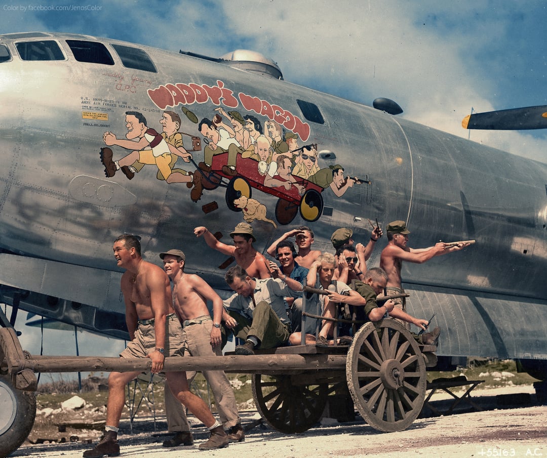 A World War II-era photo shows shirtless soldiers pulling a cart in front of a bomber plane, which features a colorful cartoon nose art of people riding in a red car with the text “Waddy’s Wagon” painted above them.