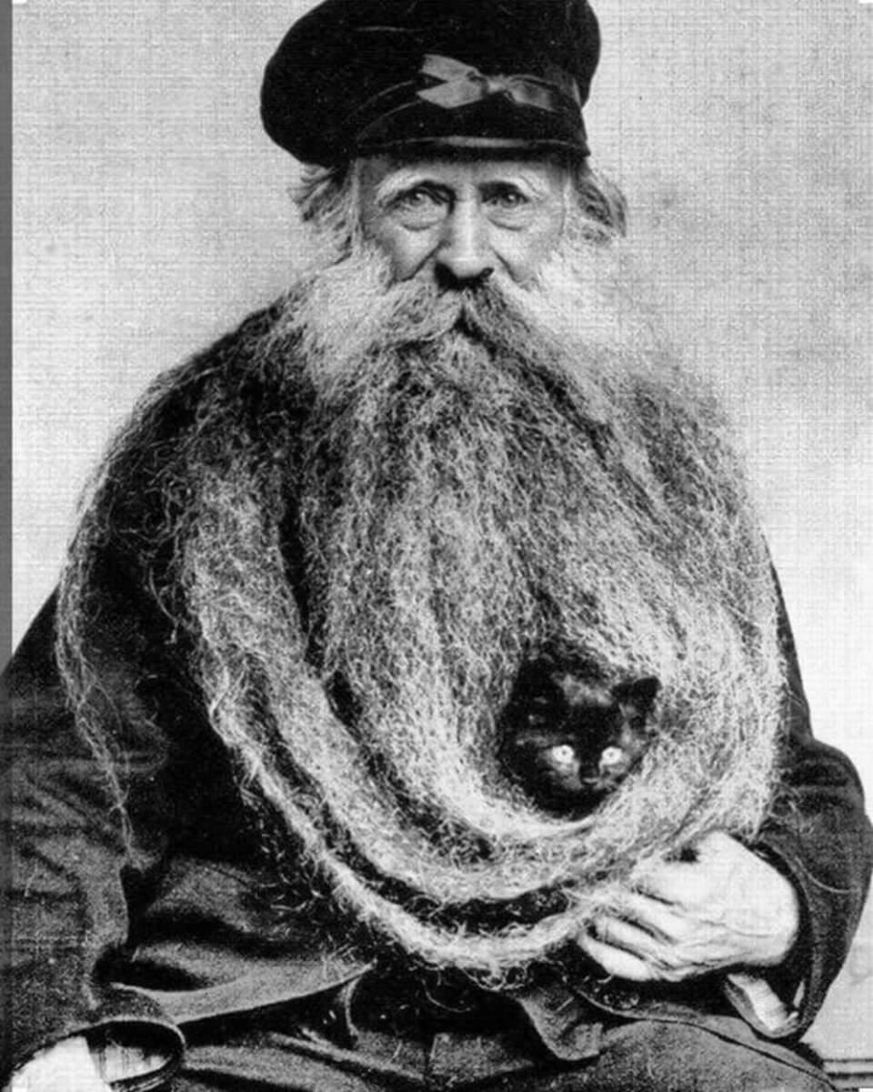 An older man with an extremely long, bushy beard sits for a portrait. A small black cat peeks out from within his beard, which is draped in loops across his chest. The man wears a dark cap and coat.
