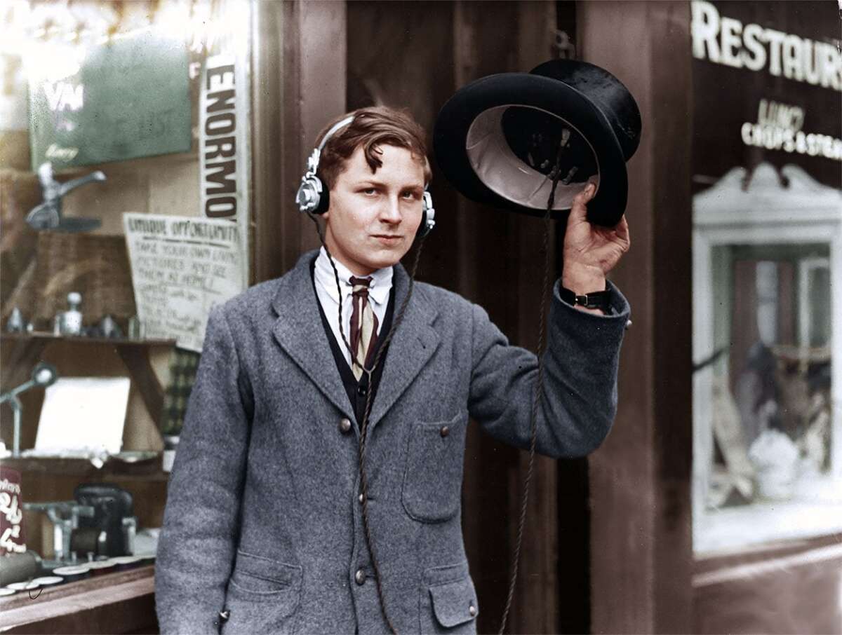 A young man in early 20th-century clothing stands outside a shop, wearing headphones and holding up a hat with wires, possibly an early portable radio device, and looks at the camera with a slight smile.