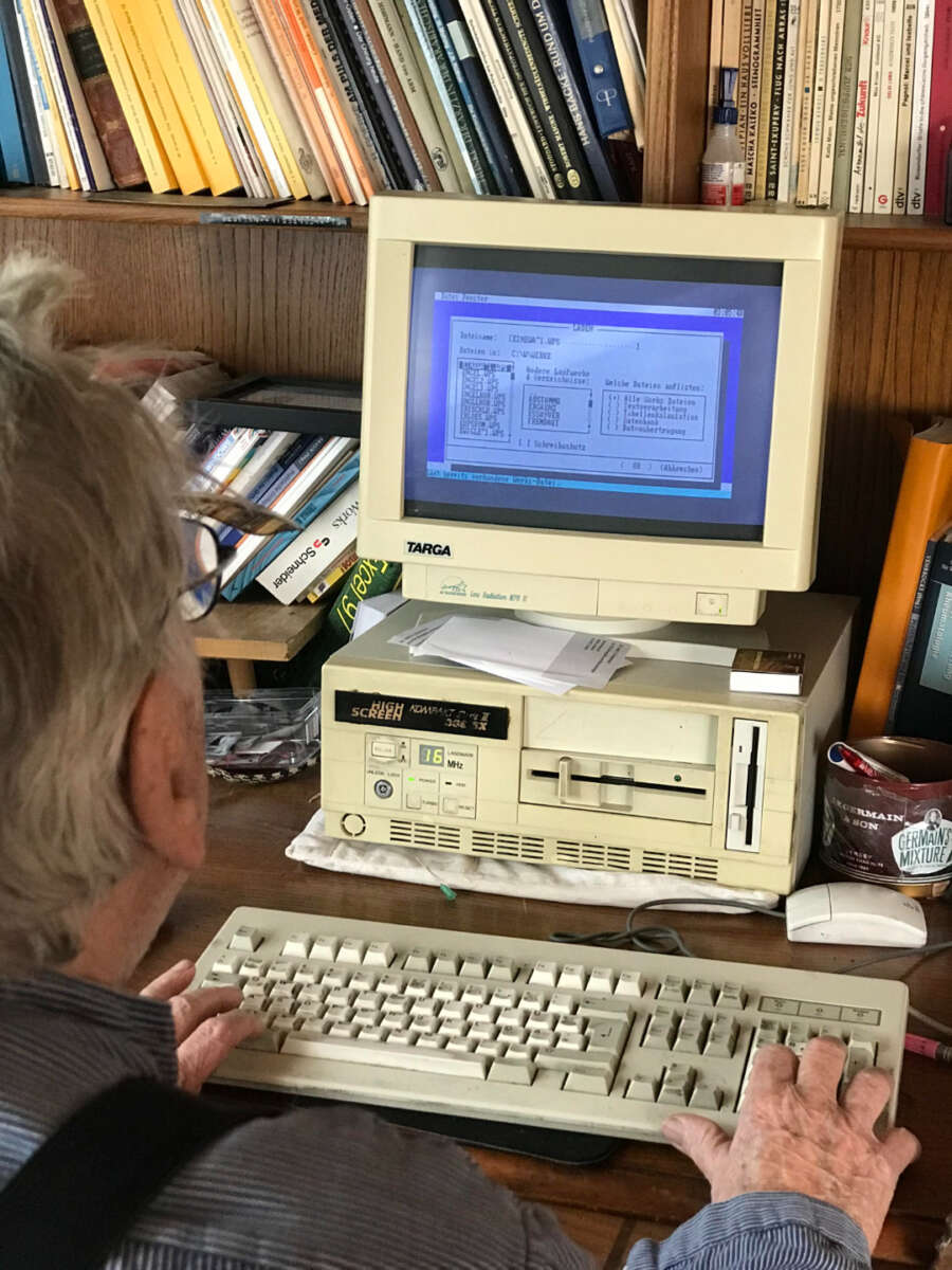 An older man types on a vintage computer with a CRT monitor displaying an old software interface, surrounded by books and papers on a wooden desk.