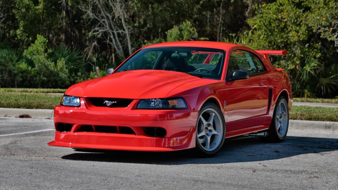 A bright red Ford Mustang coupe with a large rear spoiler is parked in an outdoor lot, surrounded by greenery and trees in the background.