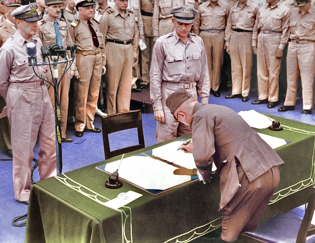 A military officer bends over a table to sign a document as uniformed officers stand behind him, witnessing the event. The scene appears formal and historic, set indoors with microphones on the table.