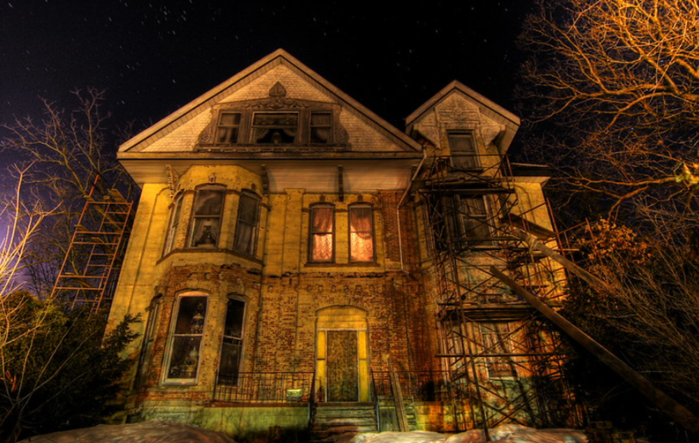 A large, old, and abandoned house at night, illuminated by eerie yellow-orange lights, with broken windows, boarded doors, and bare trees surrounding it, creating a spooky atmosphere.