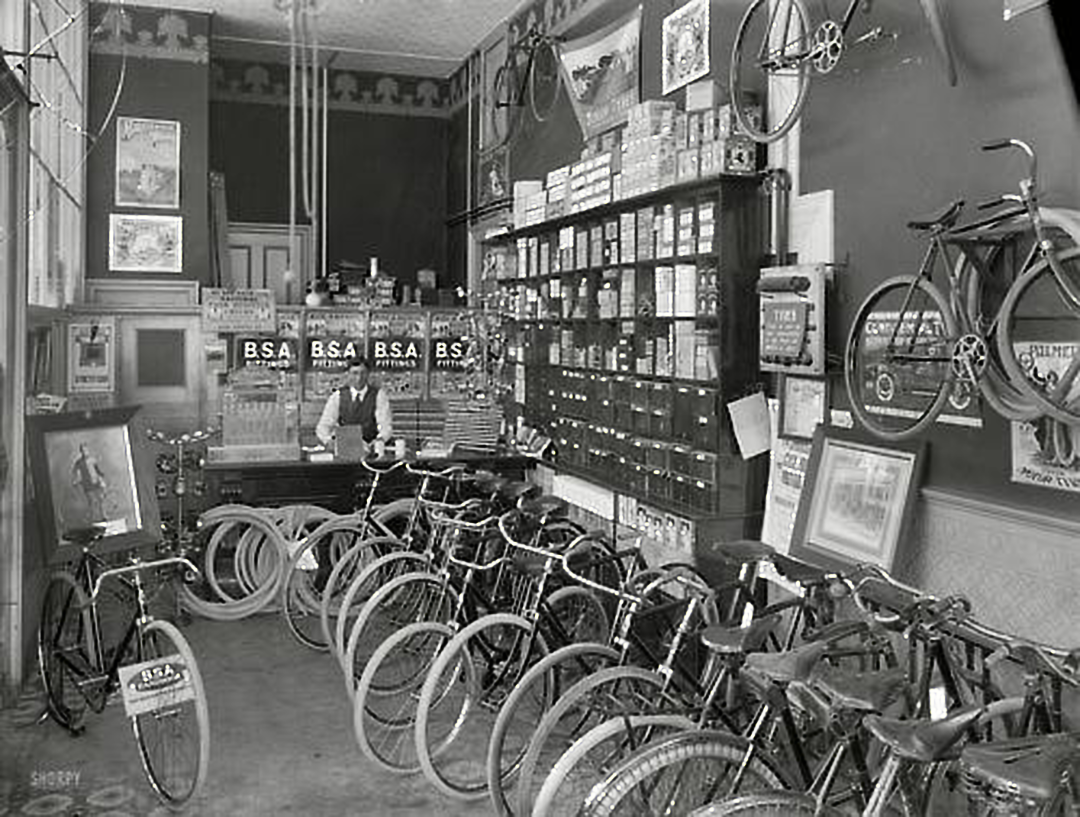 Black and white photo of a vintage bicycle shop with rows of bicycles, spare tires, posters, and shelves filled with parts. A person stands behind the counter, surrounded by tools and cycling accessories.