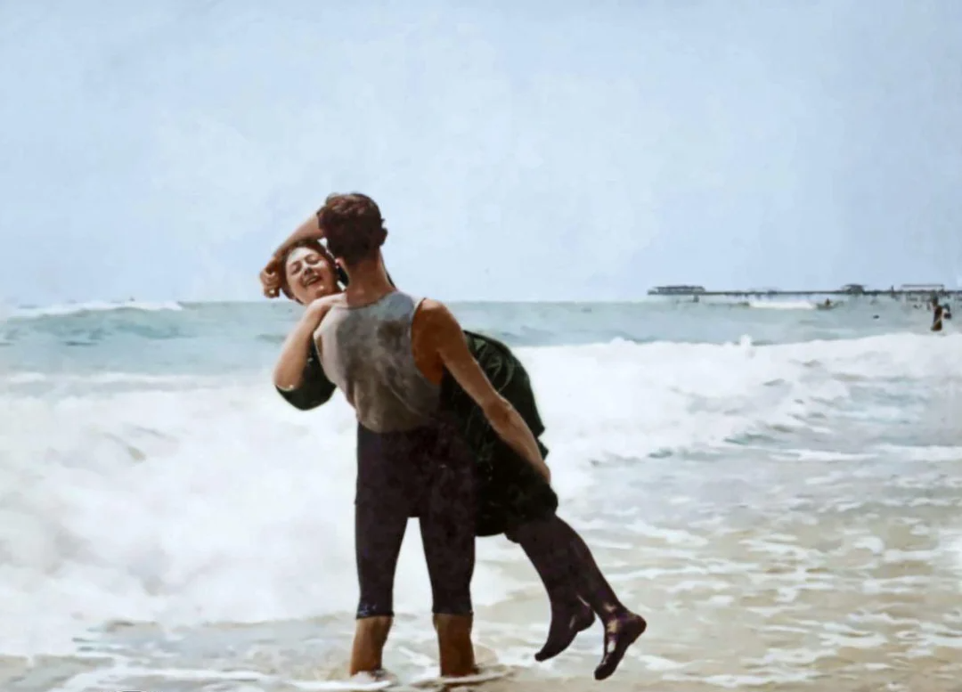 A vintage photo of a man standing in the ocean, lifting a smiling woman in his arms. Both are wearing early 20th-century bathing suits. Waves crash around them, and a pier is visible in the background.