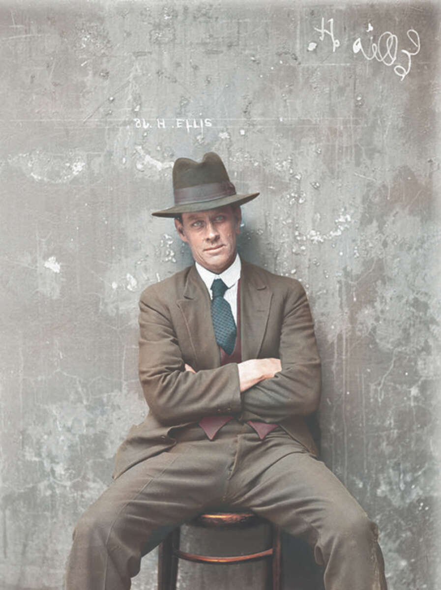 A man in a brown suit, tie, and fedora hat sits on a stool with arms folded and legs spread. He is in front of a worn, gray concrete wall with faint white markings and "H. ELLIS" written on it.