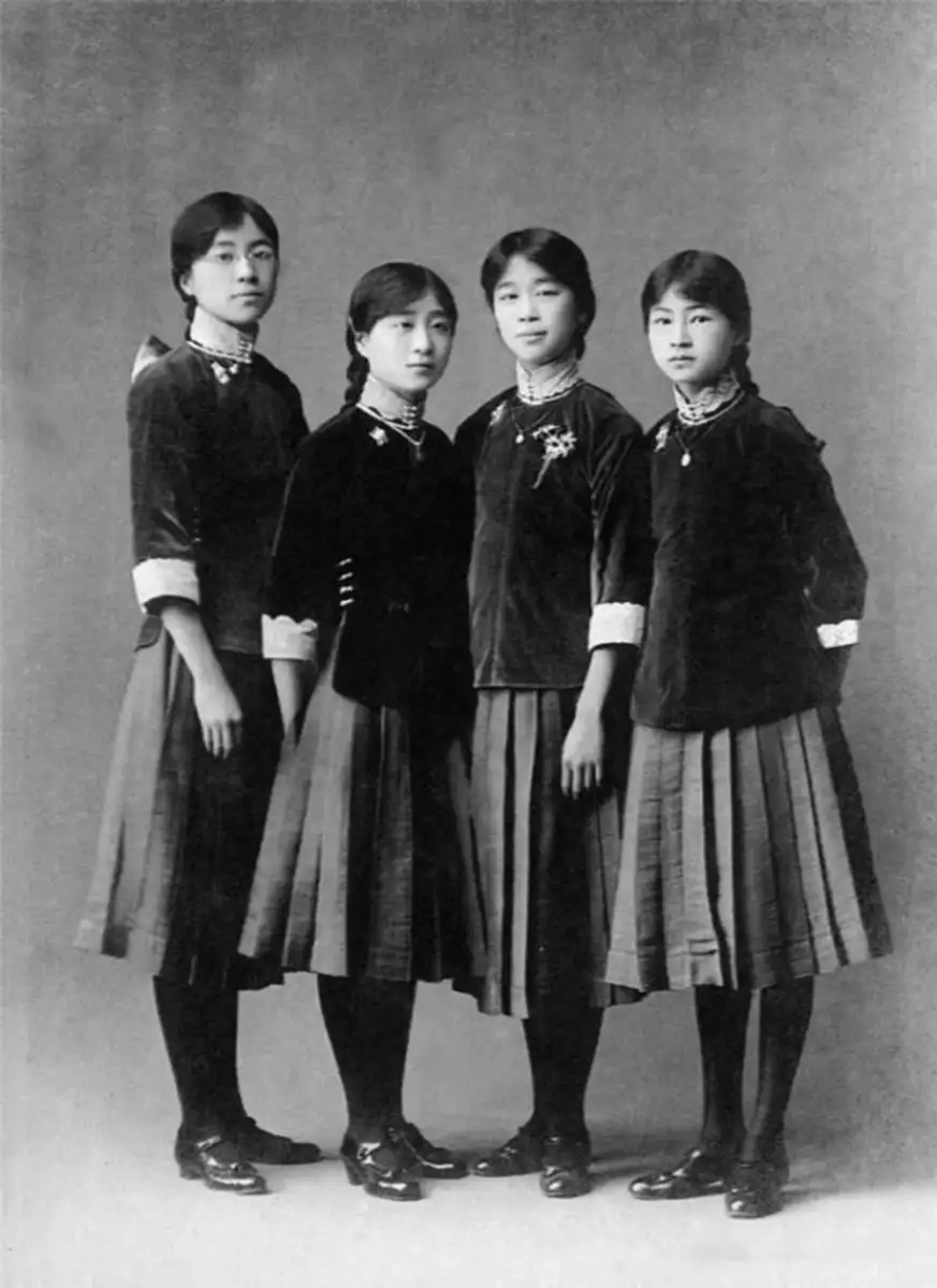 Four young women stand side by side in traditional clothing with pleated skirts and long-sleeved tops, facing the camera against a plain studio backdrop in a black-and-white vintage photograph.