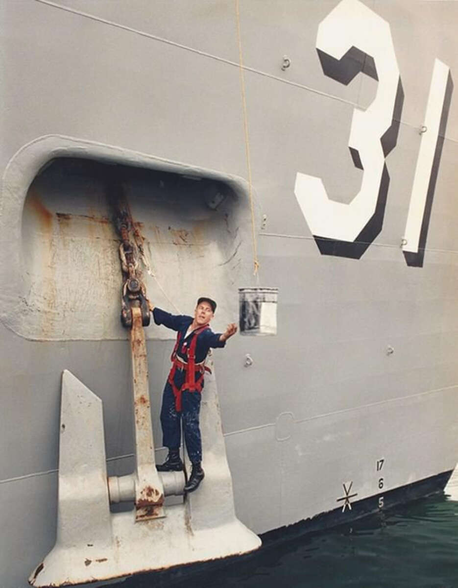 A person in a safety harness stands on a ship's anchor housing, holding onto the chain. The ship's gray hull is marked with the large white number "31" above them. Water is visible below.