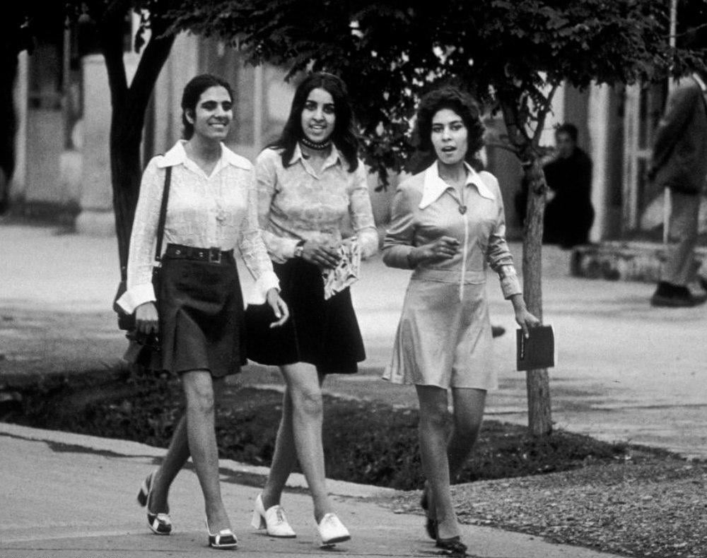 Three young women walk together on a sidewalk, smiling. They wear 1970s-style outfits with skirts and collared shirts. Trees and buildings are visible in the background.
