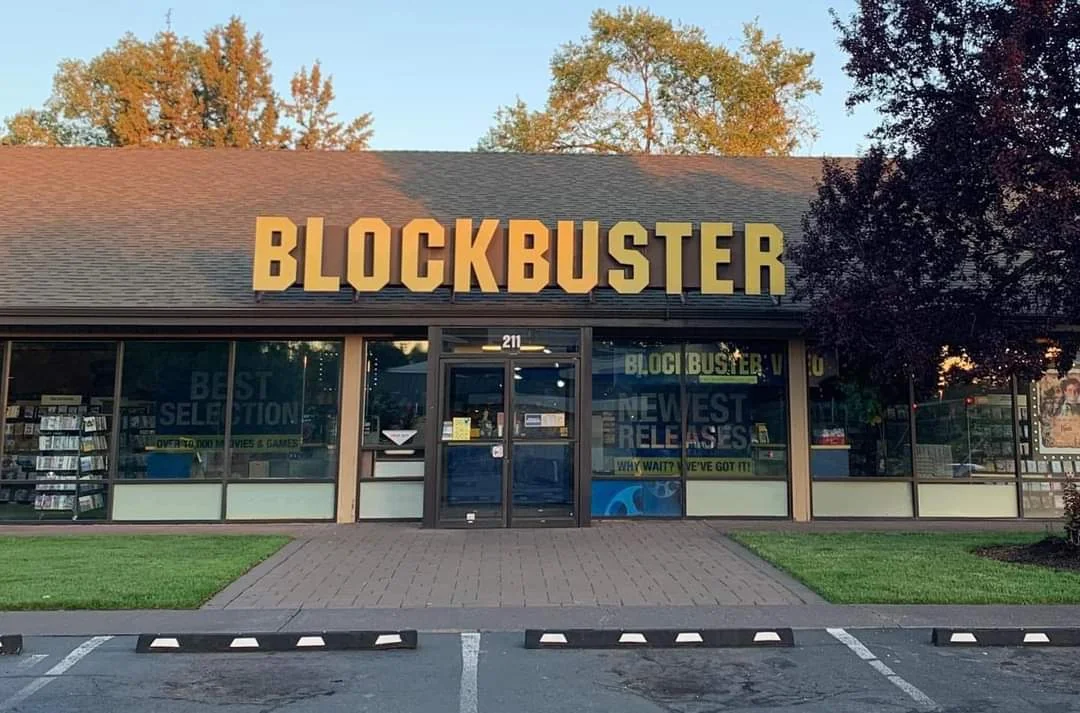A Blockbuster video rental store with large yellow letters on the storefront, surrounded by trees and a small lawn, viewed from a parking lot in front.