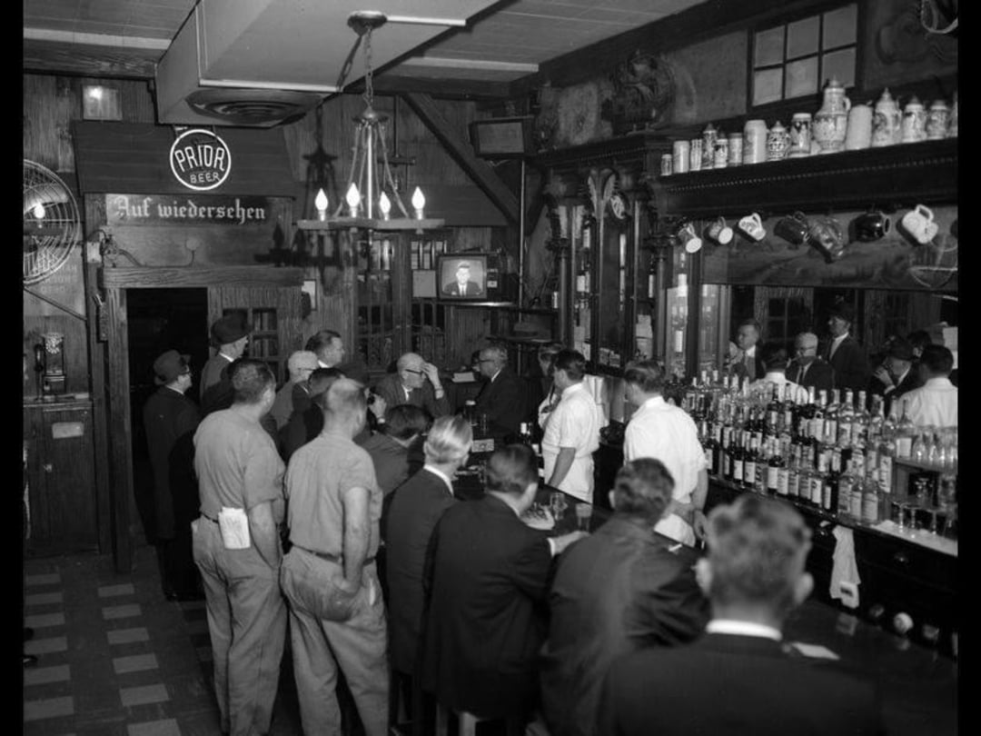 Black-and-white photo of a crowded bar with men in suits and uniforms standing or sitting at the counter. Bottles line the bar, and mirrors reflect patrons. A sign reads "Auf Wiedersehen." The setting appears vintage, likely mid-20th century.