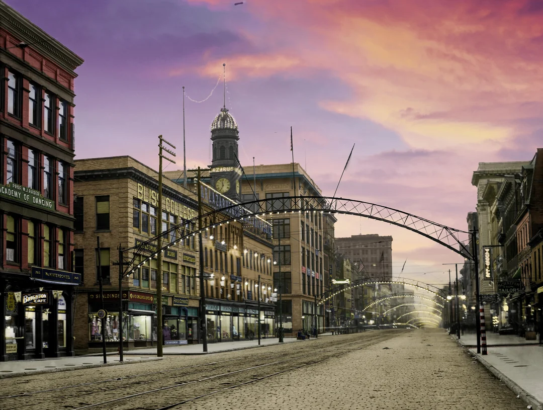 A vintage city street scene at sunset, featuring old brick buildings, arched metal streetlights, tram tracks, shop signs, and a clock tower in the background under a colorful purple and orange sky.
