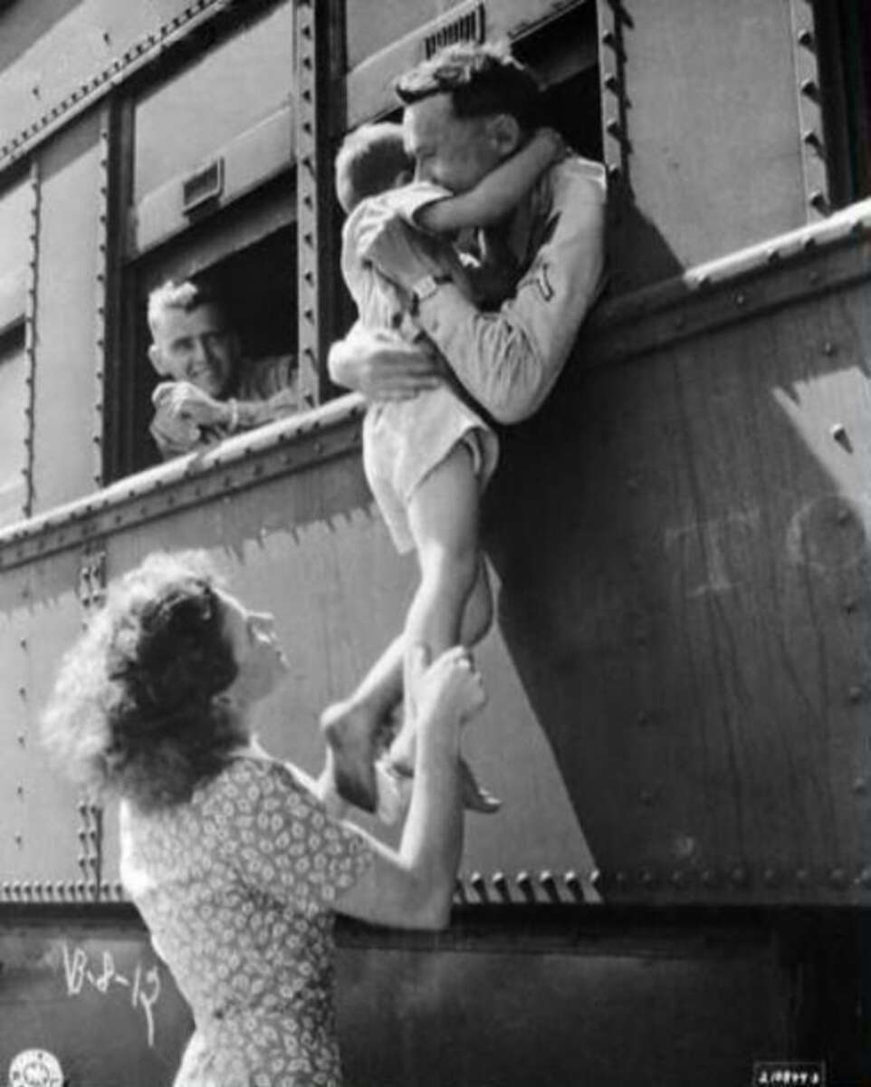 A woman lifts a smiling child up to a man leaning out of a train window for an embrace, while another man watches and smiles from a nearby window. The scene appears joyful and heartfelt.