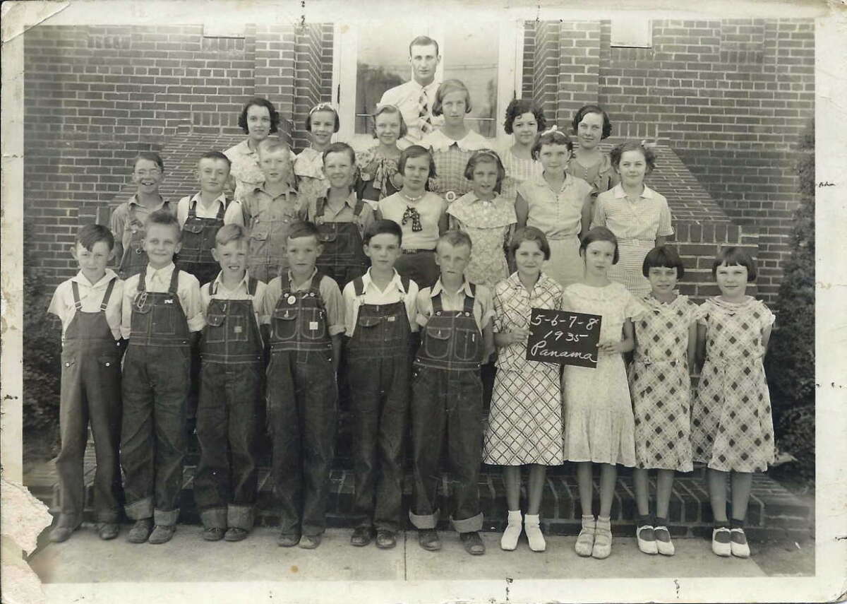 A black-and-white photo of a school class posing on brick steps. A man and several women stand behind four rows of boys and girls, some in overalls and dresses. One student holds a sign reading: "5-6-7-8 1938 Panama.