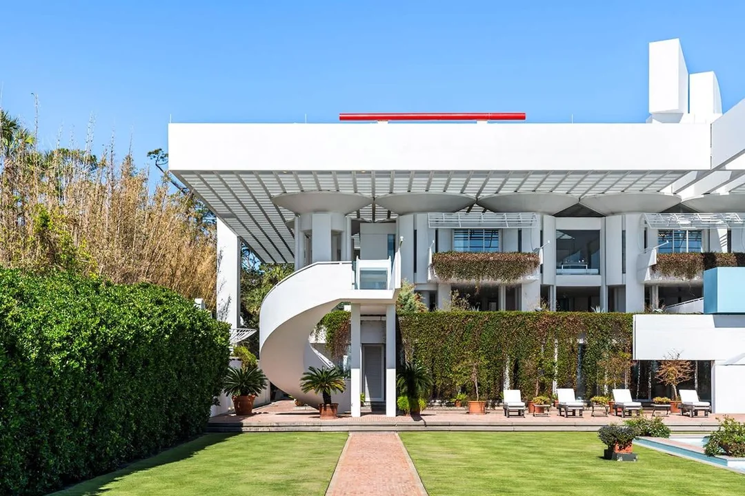 Modern white building with cylindrical columns, a spiral staircase, and balconies decorated with greenery. Manicured lawn, potted plants, and lounge chairs are visible in the foreground under a clear blue sky.