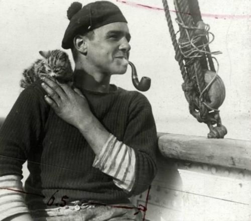 A sailor wearing a knit cap and striped shirt sits on a boat, smoking a pipe and holding a small cat on his shoulder. Rigging and a coiled rope are visible in the background. The image is black and white.