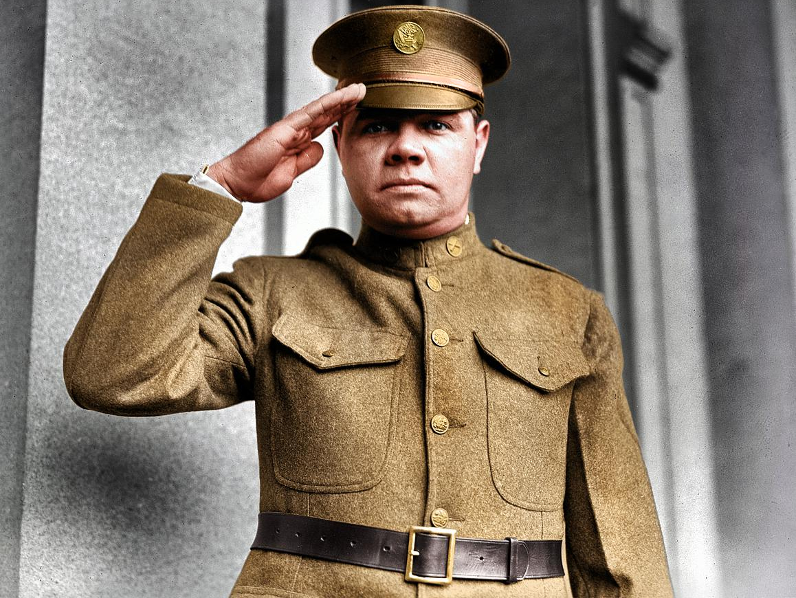 A man in a vintage military uniform stands outdoors, giving a salute. He wears a peaked cap and a double-breasted, buttoned jacket with a belt, looking directly at the camera. The background shows a building with columns.
