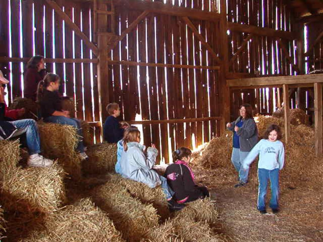 Children sit on hay bales inside a wooden barn, listening to an adult who is standing and speaking. Sunlight streams through the gaps in the wooden walls, creating a warm, rustic atmosphere. One child stands in the foreground.
