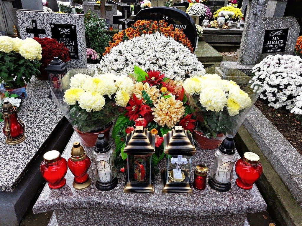 A decorated grave in a cemetery with vibrant flower arrangements, candles, and lanterns placed on and around the tombstone. White, yellow, and red flowers and several red and white votive candles are visible.