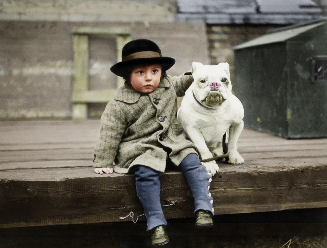 A young child in an old-fashioned coat and hat sits on a wooden platform, resting one arm around a white bulldog. Both look directly at the camera with serious expressions.